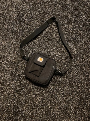 Svart axelväska från Carhartt - Säljer en svart axelväska från Carhartt med justerbar rem, fyrkantig form och flera fack framtill, inklusive ett med dragkedja och ett med lock. Perfekt storlek för det viktigaste och enkel att bära crossbody.