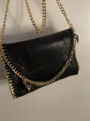 Svart axelväska med guldkedja - Säljer en svart axelväska i skinnimitation med snygga guldfärgade kedjor och dekorativ flätad kant. Väskan har ett innerfack med dragkedja och ett coolt mönstrat foder. Perfekt storlek för det viktigaste och ger en trendig touch till din outfit.