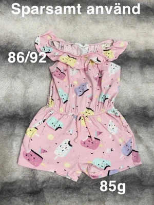 Rosa byxdress med glassmönster 86/92 - Söt rosa byxdress med färgglatt glassmönster och volang vid axlarna. Dragsko i midjan för skön passform. Perfekt för varma dagar och lek. 🌸 Sparsamt använd. Storlek 86/92
