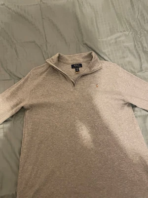 Grå half zip tröja från Polo Ralph Lauren - Säljer en stilren grå långärmad tröja från Polo Ralph Lauren med klassisk half zip och liten broderad logga på bröstet. Tröjan har en mjuk bomullskänsla och passar perfekt till lager på lager under höst och vinter.