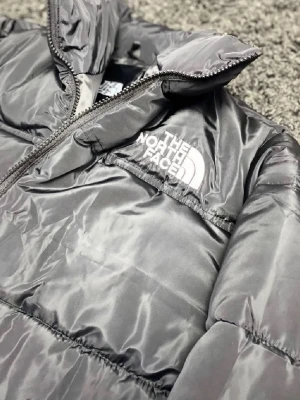 Svart pufferjacka från The North Face - Säljer en grå pufferjacka från The North Face med hög krage och dragkedja framtill. Jackan har en clean look med logga på bröstet och är vadderad för extra värme. Perfekt för kalla dagar och riktigt snygg till streetwear-stilen.