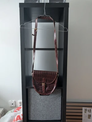 Brun axelväska i skinn med krokoprägling - Säljer en brun axelväska med krokopräglat mönster och justerbar rem. Väskan har ett snyggt spänne framtill och är tillverkad i skinn. Perfekt storlek för det viktigaste och ger en cool vibe till din outfit. Står "Made in Italy"