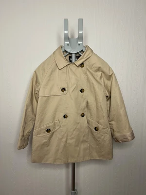 Beige trenchcoat från Zara, stl 128 - Snygg beige trenchcoat från Zara Outerwear Division med klassisk krage och dubbelknäppning. Jackan har två fickor med knappar och är tillverkad i vattenavvisande bomullsmaterial. Perfekt för regniga dagar och har en stilren look som funkar till många outfits.
