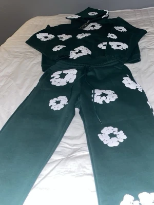 Gröna mjukisbyxor från Denim Tears - Säljer ett par gröna mjukisbyxor från Denim Tears med stora vita blommönster tryckta över hela byxorna. Byxorna har snörning i midjan och är gjorda i mjukt bomullsmaterial. Perfekt för chill dagar och streetwear vibes.