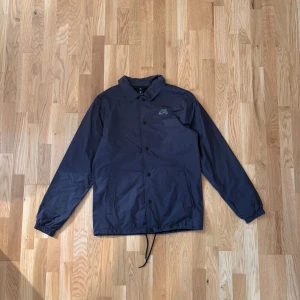 Svart vindjacka från Nike SB - Säljer en stilren svart vindjacka från Nike SB med klassisk krage och knappar framtill. Jackan har två sidofickor, snörning i nederkant och Nike SB-logga på bröstet. Perfekt för dig som gillar enkel och sportig stil.