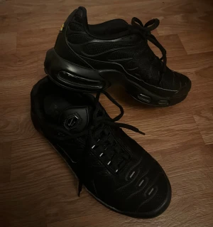 Nike Air Max TN svarta skor - Nike Air Max Plus sneakers i helsvart med TN-detalj på hälen. Snygg vågig design på ovandelen, mesh och syntetmaterial, synlig Air Max-dämpning i sulan och snörning framtill. Rund tå och platt sula för skön komfort. Perfekt för dig som gillar streetwear.