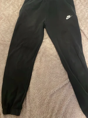 Svarta mjukisbyxor från Nike M - Svarta mjukisbyxor från Nike i storlek M. Byxorna har resår i midjan, ribbade benslut och en bakficka. Klassisk Nike-logga broderad på ena benet. Perfekta för chill eller träning.