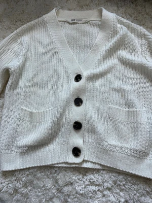 Vit stickad kofta från H&M - Säljer en vit stickad kofta från H&M i storlek 134/140. Koftan har grovstickad struktur, svarta knappar framtill, två fickor och ribbade muddar vid ärmslut och nederkant. Perfekt för lager-på-lager under kyliga dagar. Passar perfekt till jeans eller kjol!❣️❣️