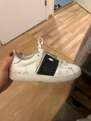 Valentino Rockstud sneakers vit/svart - Säljer ett par vita Valentino Rockstud sneakers med svart läderdetalj och svarta nitar över snörningen. Skorna har klassisk rund tå, platt sula och är tillverkade i skinn. Perfekta för dig som vill ha en edgy och lyxig touch till din outfit.