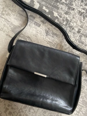 Svart axelväska i skinn  - Stilren svart axelväska från Adax i äkta skinn. Väskan har ett klassiskt, fyrkantigt format med silverfärgad detalj på locket och flera praktiska innerfack med dragkedja. Perfekt för dig som vill ha en enkel men snygg väska till vardagen.