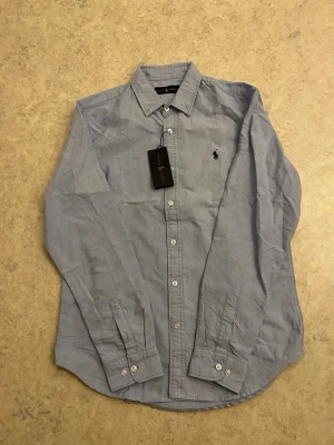 Ljusblå Ralph Lauren Skjorta M - Klassisk ljusblå skjorta från Ralph Lauren med broderad logga på bröstet. Skjortan har lång ärm, knappar hela vägen och en snygg krage. Tillverkad i mjuk bomull som känns skön mot huden. Perfekt för dig som gillar stilrena och tidlösa plagg.