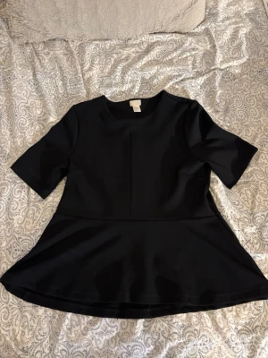 Svart peplum topp från H&M - Säljer en svart peplum topp från H&M i storlek 42. Toppen har rund halsringning, korta ärmar och en utsvängd nederdel som ger en snygg siluett. Materialet är lite tjockare och strukturerat, vilket gör att toppen sitter fint på kroppen.