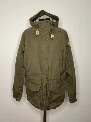 G-1000 klassisk modell från Fjällräven M  - Säljer en grön parkajacka från Fjällräven i storlek M. Jackan har rutigt fleecefoder i beige, grått och vitt, stora fickor framtill, dragsko i midjan och huva. Stängs med både dragkedja och knappar. Perfekt för kyliga dagar. OBS: Dragkedjan trasig se bild