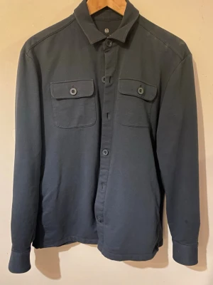 Mörkblå overshirt från Massimo Dutti - Stilren mörkblå overshirt från Massimo Dutti med klassisk krage och två bröstfickor med knappar. Tillverkad i mjuk bomull och har långa ärmar med knapp vid manschetten. Perfekt lager-på-lager-plagg för en clean look.