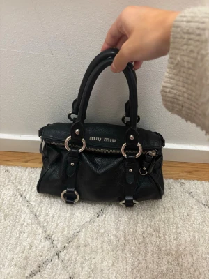 Miu Miu bow bag - Säljer den trendiga och sällsynta bow bagen ifrån miu miu. Det är en svart handväska i äkta skinn med dubbla handtag,silverfärgade detaljer runt hela väskan och rosetter på sidorna.💕Perfekt storlek för det viktigaste och riktigt snygg design, dessutom med ett fack inuti! Väskan är i super skick, däremot finns det en liten fläck på insidan, men som tur aldrig syns😊                              Storlek:Small Mått:ca 15x25