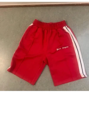 Röda Palm Angels shorts med ränder - Säljer ett par röda shorts från Palm Angels med vita och svarta ränder längs sidorna. De har elastisk midja och fickor på sidorna. Perfekta för en sportig och avslappnad stil.