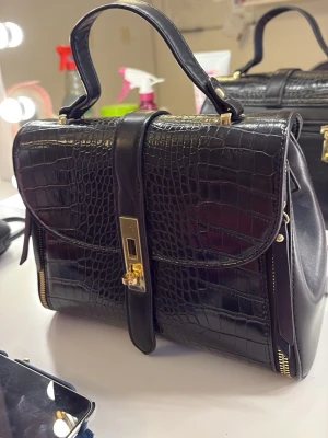 Svart handväska i krokopräglat skinn - Snygg svart handväska i krokopräglat skinn med guldfärgade metalldetaljer och elegant lås framtill. Väskan har ett stadigt handtag och dragkedjor på sidorna för extra stilpoäng. Perfekt accessoar för att lyfta din outfit. Finns axelrem till. ALDRIG ANVÄNT