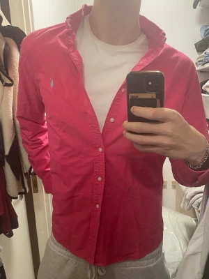 Rosa skjorta från Ralph Lauren - Säljer en snygg rosa skjorta från Ralph Lauren med klassisk krage och knappar framtill. Skjortan har det ikoniska broderade logotypen på bröstet och är tillverkad i mjuk bomull. Perfekt för dig som vill sticka ut med färg och stil.