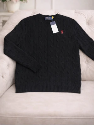 Svart kabelstickad tröja Polo Ralph Lauren - Kabelstickad svart tröja från Polo Ralph Lauren med klassisk röd broderad logga på bröstet. Rund halsringning och ribbade muddar vid ärmslut och nederkant. Tillverkad i mjuk bomull som känns skön mot huden.
