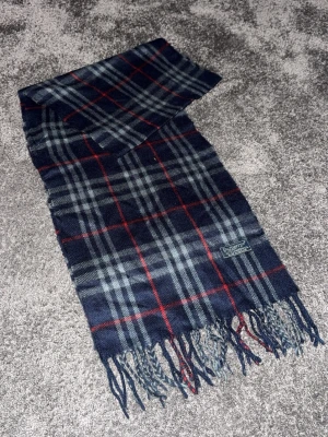 Rutig lammullhalsduk från Burberry - Säljer en klassisk rutig halsduk från Burberry i marinblått, grått och rött. Halsduken är tillverkad i 100% lammull och har fransar i ändarna. Perfekt accessoar för att lyfta din outfit med en tidlös brittisk touch.