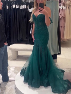 Grön balklänning med spetsdetaljer - Säljer en smaragdgrön långklänning med offshoulder-ärmar och markerad midja. Klänningen har en figurnära passform upptill och vid kjol med släp. Tyll och spetsdetaljer ger en lyxig känsla, perfekt för bal eller fest. Hjärtformad urringning och korsett upptill. Klänningen är limited edition så går inte längre att köpa! Prislappen är kvar och endast testad