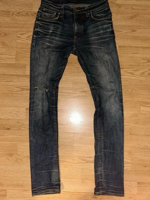 Mörkblå jeans från Nudie Jeans - Säljer ett par mörkblå jeans från Nudie Jeans med snygga slitningar och kontrastsömmar. Klassisk femficksmodell med orange detaljer och broderade bakfickor. Jeansen har ett hål vid knät för att ge en snygg detalj. Hör av dig vid intresse. MVH 