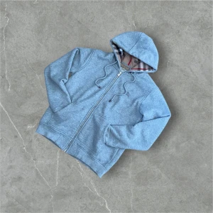 Grå hoodie med dragkedja från Burberry - Säljer en grå hoodie från Burberry med dragkedja och klassiskt rutigt foder i huvan. Jackan har snörning i huvan och broderad logga på bröstet. Perfekt för en avslappnad stil.