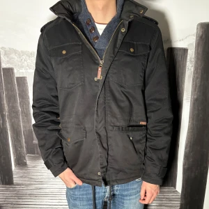 Svart parkajacka med dragkedja - Säljer en svart parkajacka med dragkedja och tryckknappar. Jackan har flera fickor framtill, axelklaffar och en klassisk krage. Perfekt för dig som vill ha en stilren och funktionell jacka till vardags. Passar till jeans och sneakers.