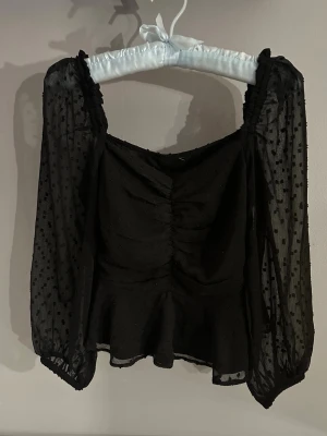 Svart blus med transparenta ärmar - Säljer en svart blus med transparenta, långärmade ärmar och små prickar i tyget. Toppen har rynkade detaljer framtill och en lätt volang vid nederkanten. Perfekt för dig som gillar en romantisk och trendig look.