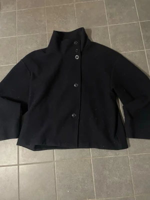 Svart ulljacka med hög krage - Snygg svart jacka i ull med hög krage och raka, vida ärmar. Jackan har knäppning med fyra knappar framtill och en minimalistisk, stilren look. Perfekt för dig som gillar enkel men trendig design.