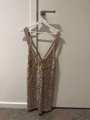 Glittrig paljettklänning - Säljer en glittrig kortklänning från NAKD i storlek EU 38. Klänningen har multifärgade paljetter, breda ljusrosa axelband och snygg knytning i ryggen. Prislapp kvar så aldrig använd!!
