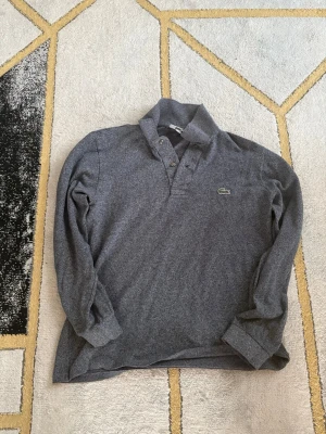 Grå långärmad pikétröja från Lacoste i enkel och stilren design. - Mörkgrå långärmad henley från Lacoste med klassisk krage och knappar framtill. Ikoniska gröna krokodilloggan på bröstet. Skön och stilren tröja i mjukt bomullsmaterial, perfekt till chill eller skola.