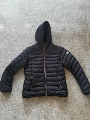 Svart Moncler dunjacka med huva - Snygg dunjacka Moncler med huva och fluffigt foder. Jackan har quiltad design, dragkedjor med röd-vita detaljer och klassisk Moncler-logga på ärmen. Perfekt för kalla dagar när du vill hålla stilen och värmen. Loggan sitter lite dåligt.