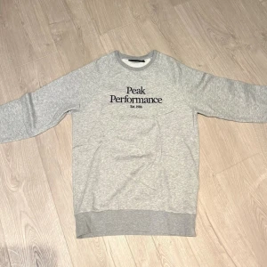 Grå sweatshirt från Peak Performance - Säljer en klassisk grå sweatshirt från Peak Performance med svart logga på bröstet. Tröjan har rund halsringning, långa ärmar och ribbade muddar vid ärmslut och nederkant. Perfekt för en avslappnad och sportig stil.