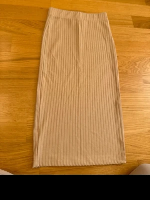 Beige ribbad midikjol från NA-KD - Säljer en beige ribbad midikjol från NA-KD. Kjolen har en tight passform och är tillverkad i ett  stretchigt material som sitter snyggt på kroppen. Perfekt att styla med en enkel topp eller hoodie.