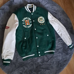 Grön och vit bomberjacka H&M Hawkins - Snygg grön och vit bomberjacka från H&M med Hawkins High School-tryck och broderad tiger på bröstet. Jackan har vita ärmar, grönt tyg på kroppen, ribbade muddar och knappar framtill. Perfekt för dig som gillar Stranger Things och college-stil.
