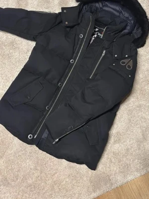 Svart Moose Knuckles dunjacka - Säljer en svart dunjacka från Moose Knuckles med avtagbar huva och fuskpäls. Jackan har flera fickor, dragkedjor och tryckknappar samt en snygg logga på ärmen. Insidan har ett broderat motiv med hockeyspelare. Perfekt för kalla dagar!