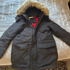 Svart Jacka Tommy Hilfiger - Svart jacka från Tommy Hilfiger med stor logga på ryggen och emblem på ärmen. Jackan har huva med fuskpäls. Använd några gånger🌸