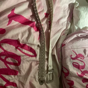 Vitt glittrigt bälte med nitar - Säljer ett vitt bälte med glittriga stenar och silvriga nitar. Bältet har en bredare form och ett coolt mönster med blanka detaljer längs hela längden. Spännet är dekorerat med stenar och ger en extra bling-effekt. Perfekt för att lyfta din outfit med lite extra edge. Fattas en sten se bild 