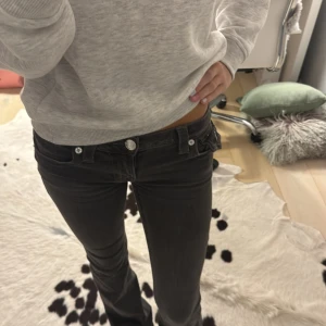 Mörkgrå true religion jeans - Snygga mörkgrå jeans med låg midja och bootcut passform. Byxorna har dekorativa fickor bak med lock och knapp, samt klassiska jeansdetaljer framtill. Perfekta för dig som gillar en trendig och avslappnad stil.