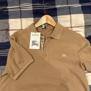 Beige pikétröja från Burberry - Snygg beige pikétröja från Burberry med klassisk krage och korta ärmar. Tröjan har Burberrys ikoniska rutor vid knappslån och broderad logga på bröstet. Tillverkad i mjuk bomull som känns skön mot huden. Perfekt för dig som gillar stilrena och tidlösa plagg. ( oanvänd )