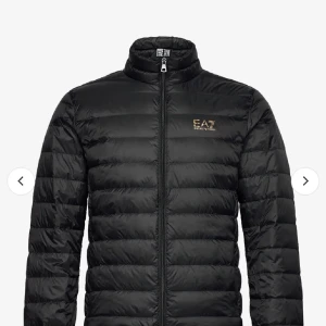 Svart pufferjacka från EA7 Armani - Svart pufferjacka från EA7 Emporio Armani med guldfärgat logotryck på bröstet. Jackan har ståkrage, dragkedja framtill och quiltad design. Perfekt för dig som vill ha en stilren och varm jacka med exklusiv känsla. Passar mer som S 