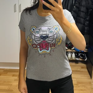 Grå Kenzo t-shirt med tigertryck - Säljer en grå t-shirt från Kenzo med ikoniskt tigertryck i flera färger och Kenzo Paris-text över bröstet. T-shirten har korta ärmar och rund hals, perfekt för dig som gillar statement-plagg och streetstyle.