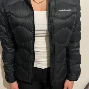Svart pufferjacka Peak Performance - Svart pufferjacka, Helium Down jacket dam från Peak Performance med snyggt vågformat quiltat mönster. Jackan har dragkedja framtill, lång ärm och diskret logga på bröstet och ärmen. Den är dunvadderad så perfekt för aktiviteter utomhus. Passformen är regular. Nypris 2800. Änvänd cirka 5 gånger så väldigt nytt skick 