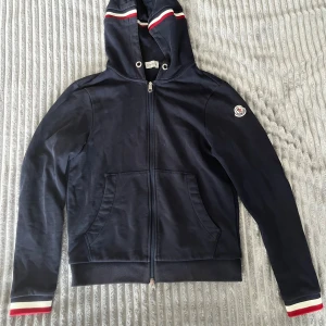 Navy blå Moncler hoodie med huva - Snygg navy blå hoodie från moncler med dragkedja, huva och klassiska röda och vita ränder på huva och även handleder. Tröjan har två fickor fram och moncler-logga på ärmen. Perfekt för dig som gillar slitrena och sportiga plagg, går att skanna så den är ÄKTA! Tveka inte på att höra av dig vid minsta fundering 🙌
