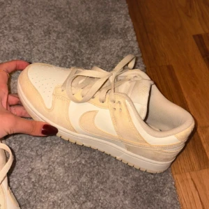 Nike Dunk Low i beige och vitt - Säljer ett par Nike Dunk Low sneakers i storlek 39. Skorna har en snygg kombination av beige mocka och vita detaljer i skinn, med klassisk Nike-logga på sidan och broderad logga bak. Perfekta för dig som gillar stilrena och neutrala färger.
