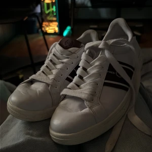 Vita Fred Perry sneakers med vinröda detaljer - Säljer ett par klassiska vita sneakers från Fred Perry med vinröda detaljer och guldlogga på hälen. Skorna har snörning, rund tå och två vinröda ränder på sidan. Tillverkade i skinn med en platt sula, perfekta för en clean och sportig stil.