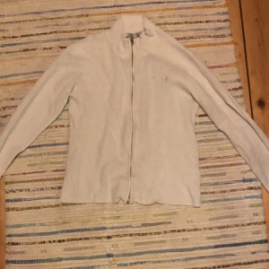 Vit ribbad kofta med dragkedja 157 - Vit ribbad kofta från 157 med hel dragkedja framtill och ståkrage. Tröjan har långärmad passform och är tillverkad i mjukt, stretchigt material som känns skönt mot huden. Perfekt för dig som gillar stilrena och enkla plagg med lite edge.