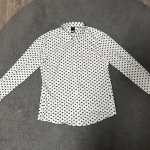 Vit blommig skjorta från BOSS XL - Snygg vit skjorta från BOSS med brunt blommigt mönster över hela tyget. Skjortan har klassisk krage, långa ärmar och knäppning framtill. Tillverkad i bomull för en skön känsla och stilren look. Nypris ligger runt 1500kr