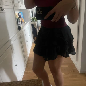 Svart volangkjol med resårmidja - Säljer en svart kortkjol med flera lager volanger och resår i midjan. Kjolen har en luftig och flowy känsla, perfekt för dig som gillar att sticka ut med en cool och feminin stil. Materialet är lätt och ger snygg rörelse när du går.